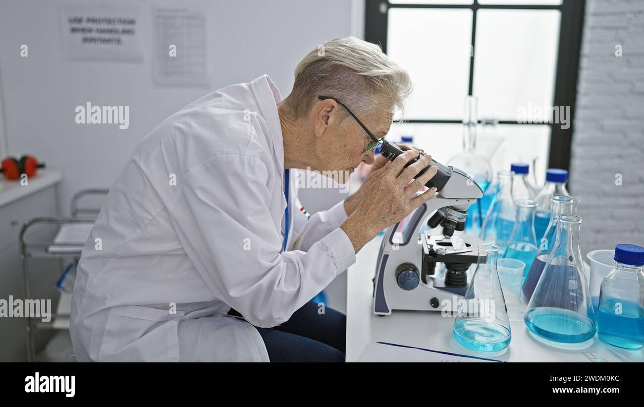 Scientifique senior dédiée aux cheveux gris immergée dans un travail intense de recherche au microscope au niveau du doctorat en laboratoire Banque D'Images