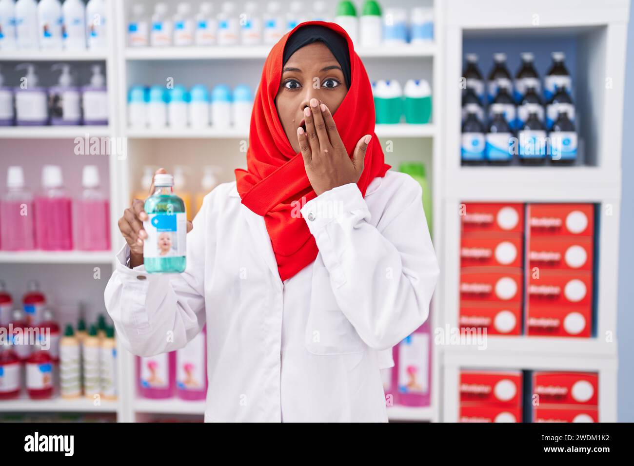 Jeune femme arabe travaillant à la pharmacie pharmacie tenant le sirop couvrant la bouche avec la main, choquée et effrayée pour erreur. expression surprise Banque D'Images