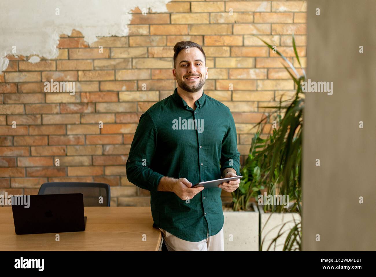Un homme joyeux tient avec confiance une tablette numérique dans un espace de bureau contemporain avec un mur de briques apparentes, symbolisant un mélange de technologie moderne Banque D'Images