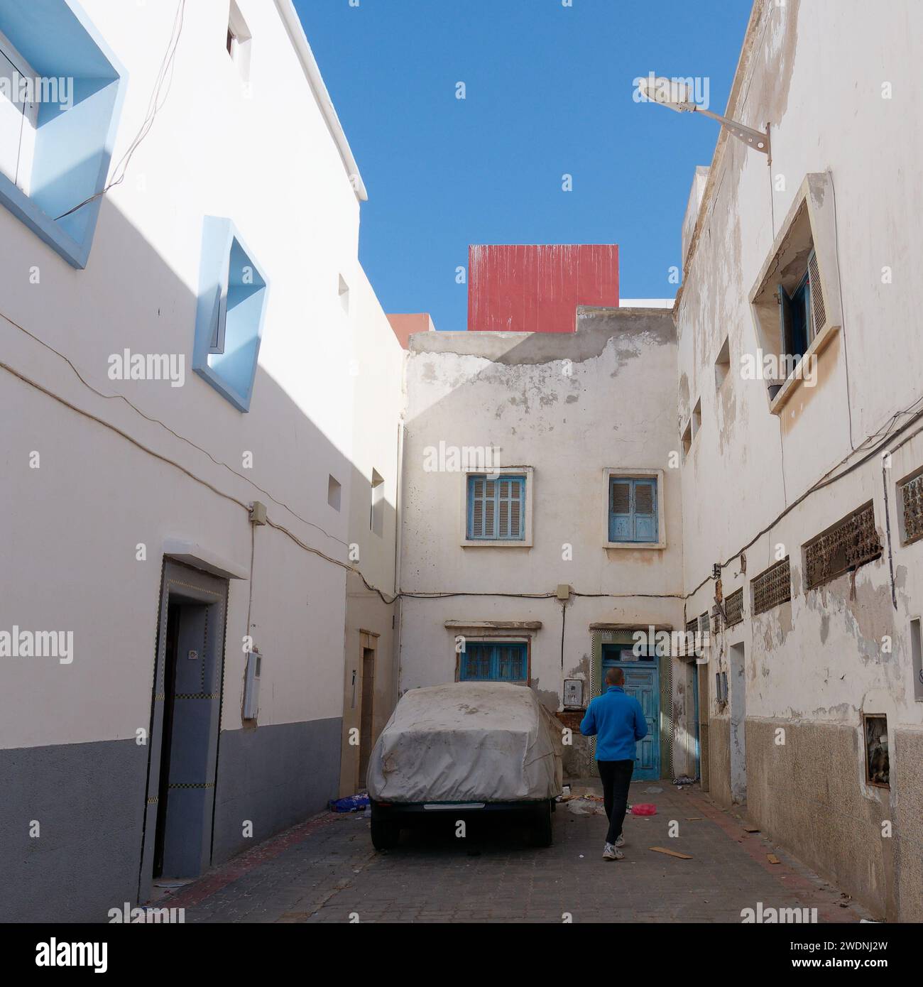 Homme en haut bleu marche dans une étroite impasse avec une voiture avec une couverture à Essaouira, Maroc, 21 janvier 2024 Banque D'Images