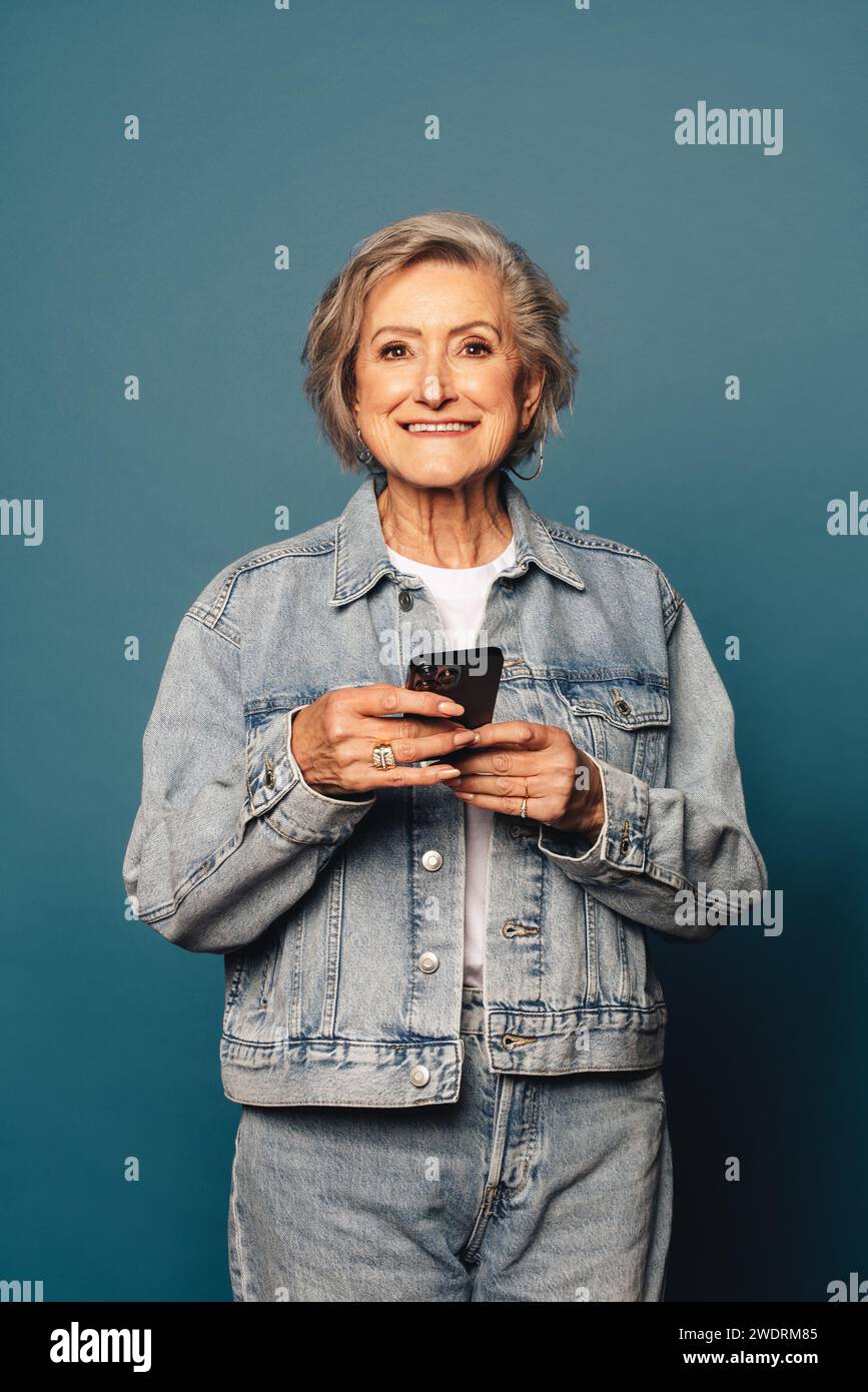 Portrait d'une femme mature en vêtements denim décontractés se tient dans un studio, tenant un smartphone. Avec un sourire joyeux, elle regarde directement la caméra Banque D'Images