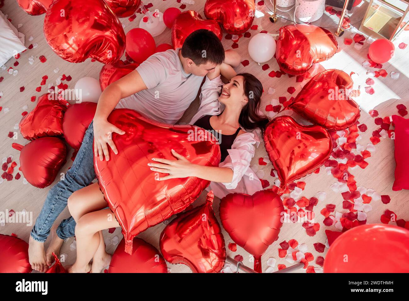 Vue de dessus d'un jeune couple gai couché sur le sol près de ballon blanc rouge en forme de coeur, pétales de rose dispersés autour. Décoration romantique Saint Valentin Banque D'Images