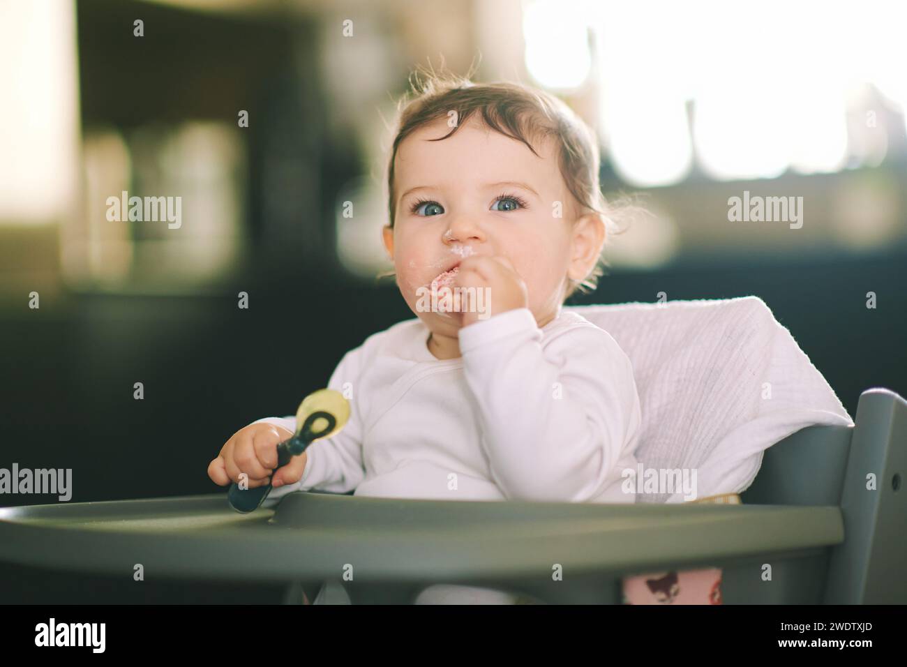 Portrait d'adorable bébé en désordre assis dans une chaise haute, tenant une cuillère Banque D'Images