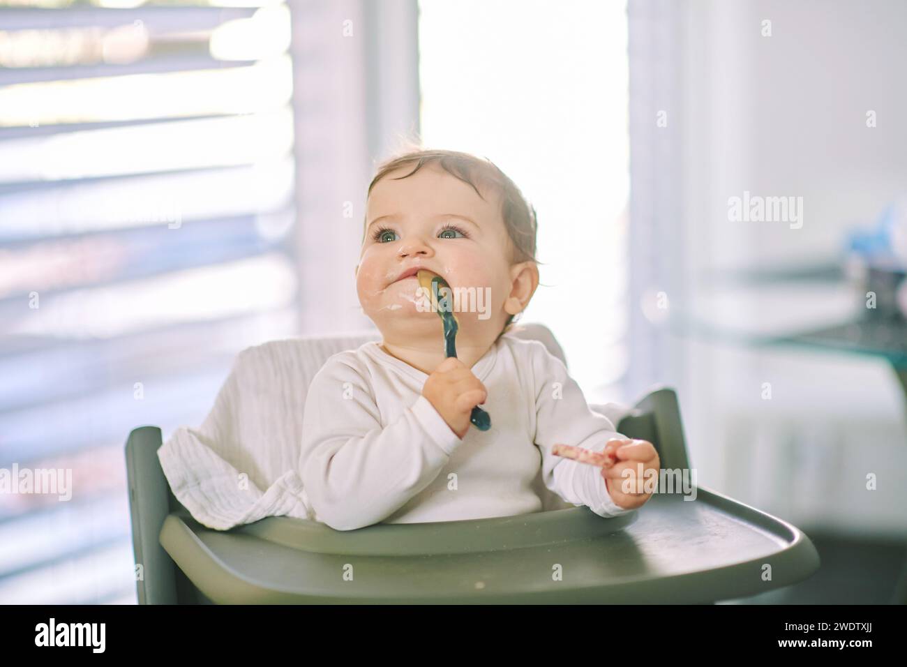 Portrait d'adorable bébé en désordre assis dans une chaise haute, tenant une cuillère Banque D'Images