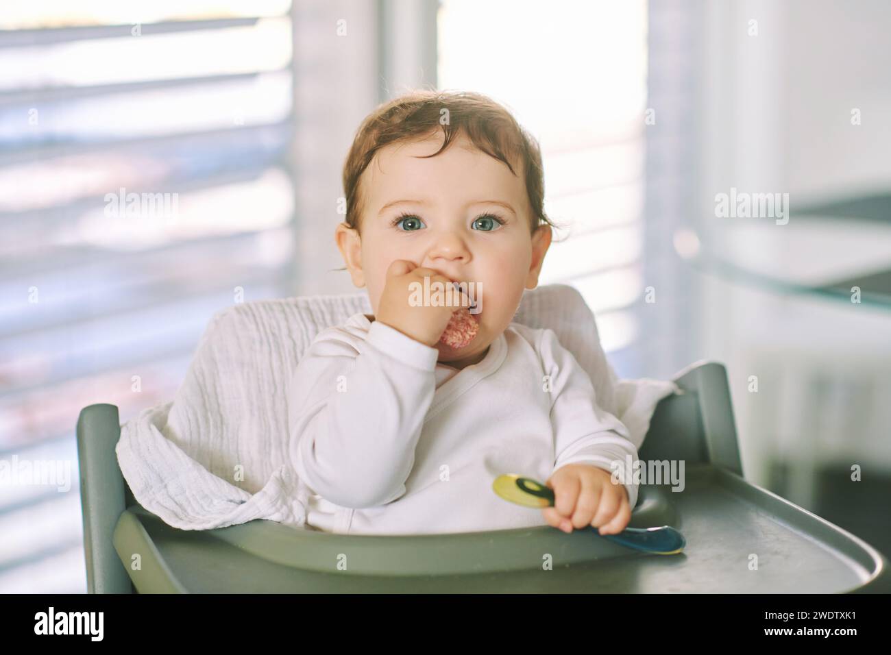 Portrait d'adorable bébé en désordre assis dans une chaise haute, tenant une cuillère Banque D'Images