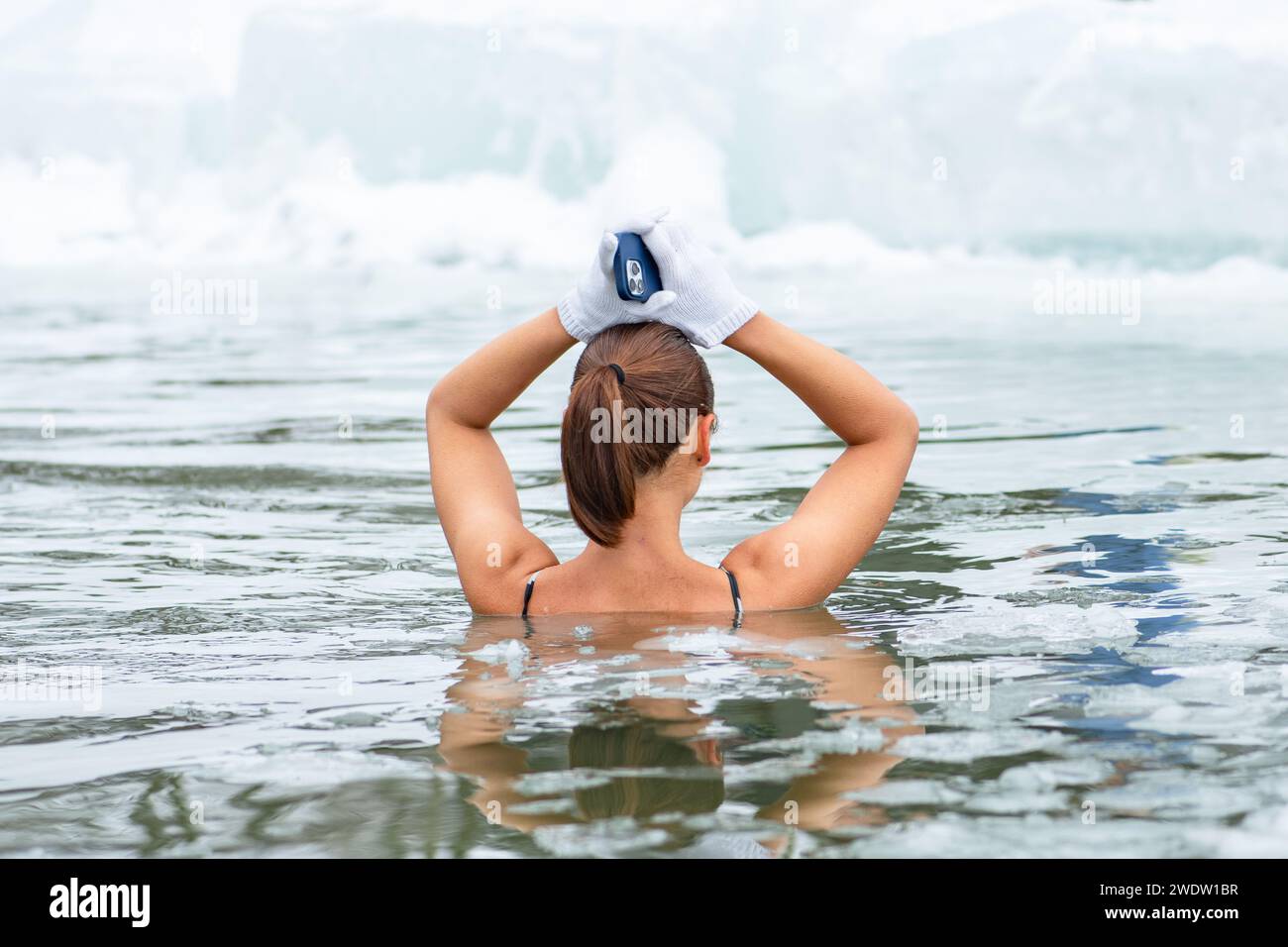 Belle fille ou femme avec un bain de glace de téléphone dans l'eau froide glaciale d'un lac gelé. Méthode Wim Hof, thérapie par le froid, techniques respiratoires, yoga Banque D'Images