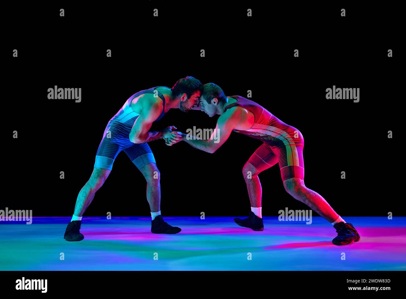 Jeune athlète homme, lutteurs en uniforme bleu et rouge luttant à la main en position neutre sur leurs pieds dans des lumières de néon sur fond noir. Banque D'Images
