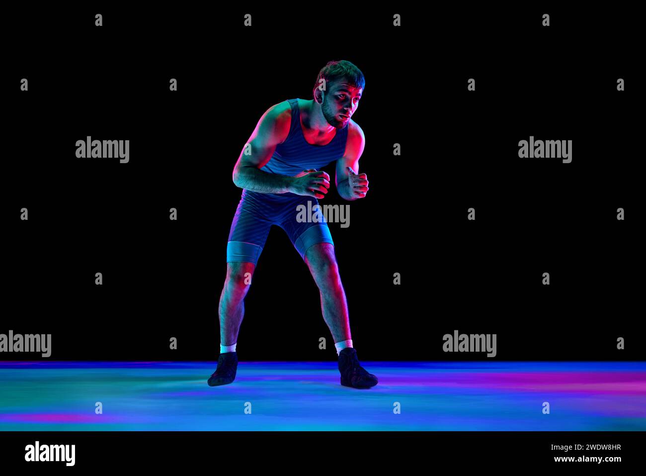 Sportsman, lutteur freestyle en collants de combat bleus se tient en position d'attaque et regarde ta caméra sur fond noir dans des lumières de néon mixtes. Banque D'Images