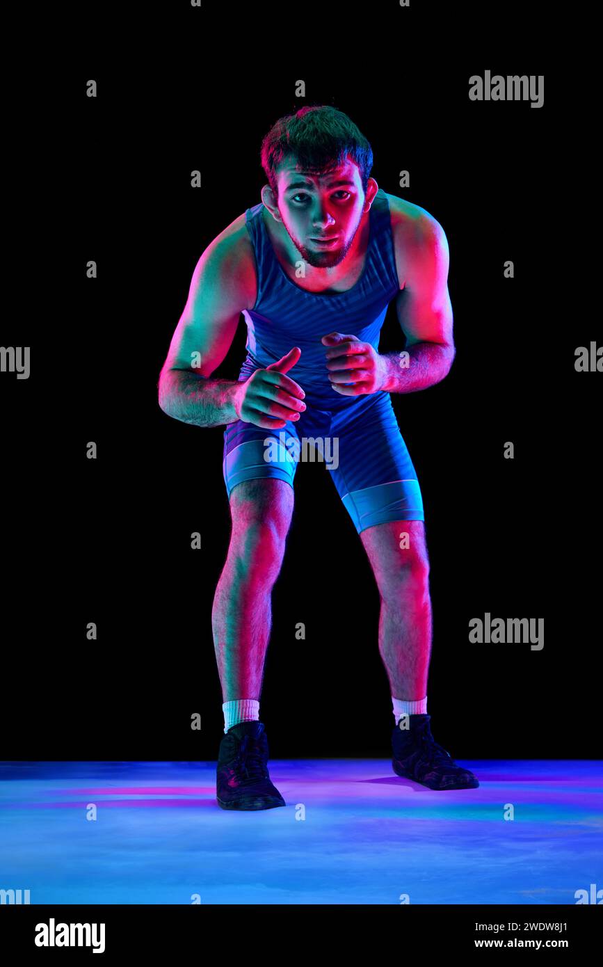 Sportsman, lutteur freestyle dans des collants de sport bleus se tient en position d'attaque et regardant ta caméra sur fond noir dans des lumières de néon mixtes. Banque D'Images