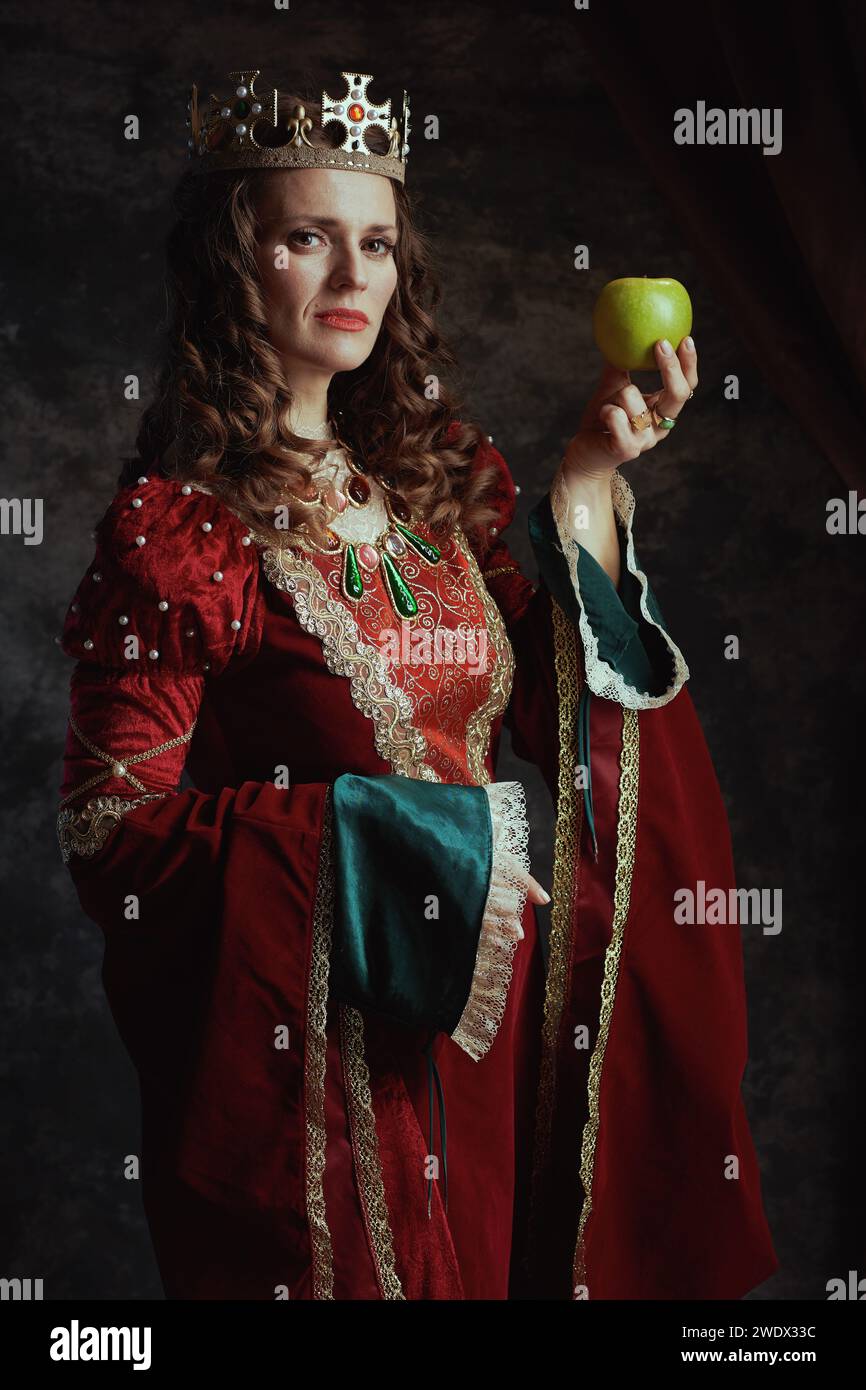 reine médiévale en robe rouge avec pomme verte et couronne sur fond gris foncé. Banque D'Images