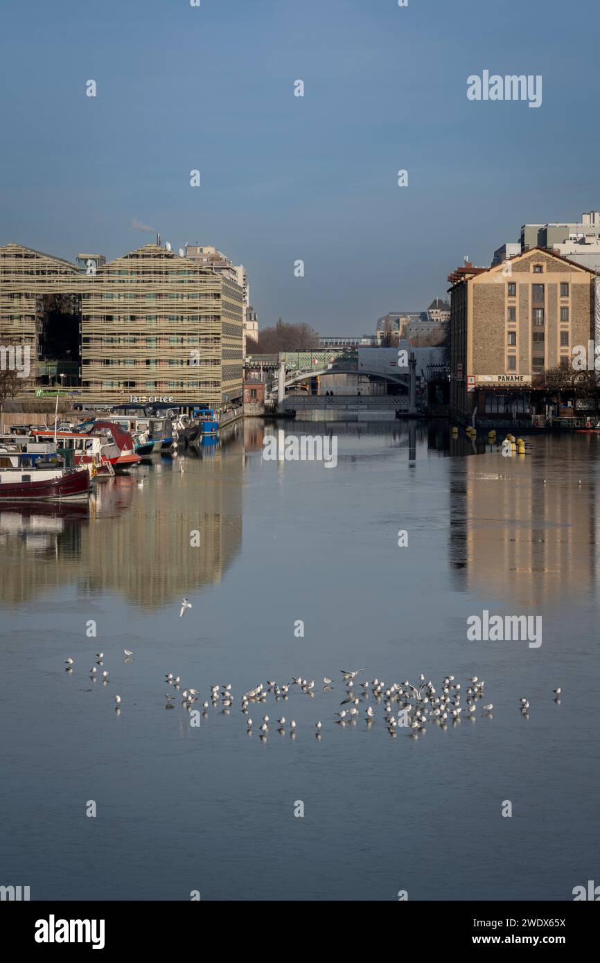 Paris, France - 01 20 2024 : Canal de l'Ourcq. Réflexions sur le canal de l'Ourcq d'immeubles résidentiels, pont, barges et mouettes sur eau gelée Banque D'Images