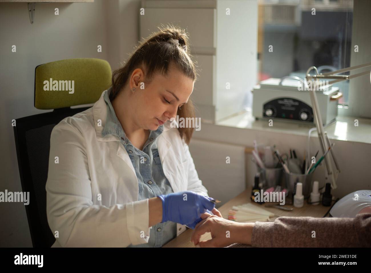 Technicienne en ongles portant des gants, un manteau blanc et est concentrée tenant la main de son client tout en travaillant Banque D'Images