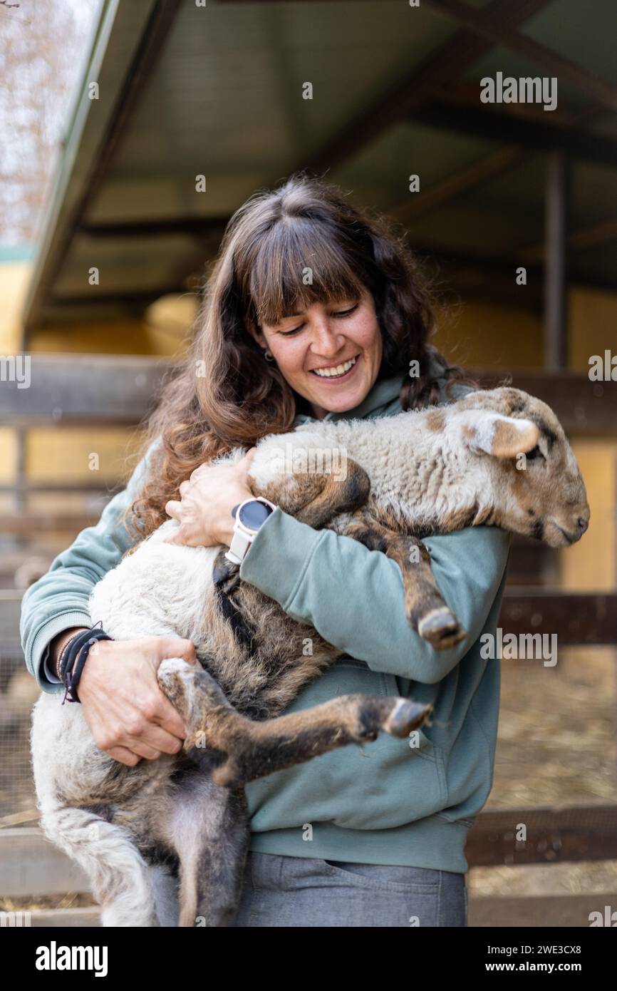 Heureuse jeune femme fermière serrant un bébé mouton sur une ferme biologique rurale. Bien-être et soins des animaux dans une grange. Banque D'Images