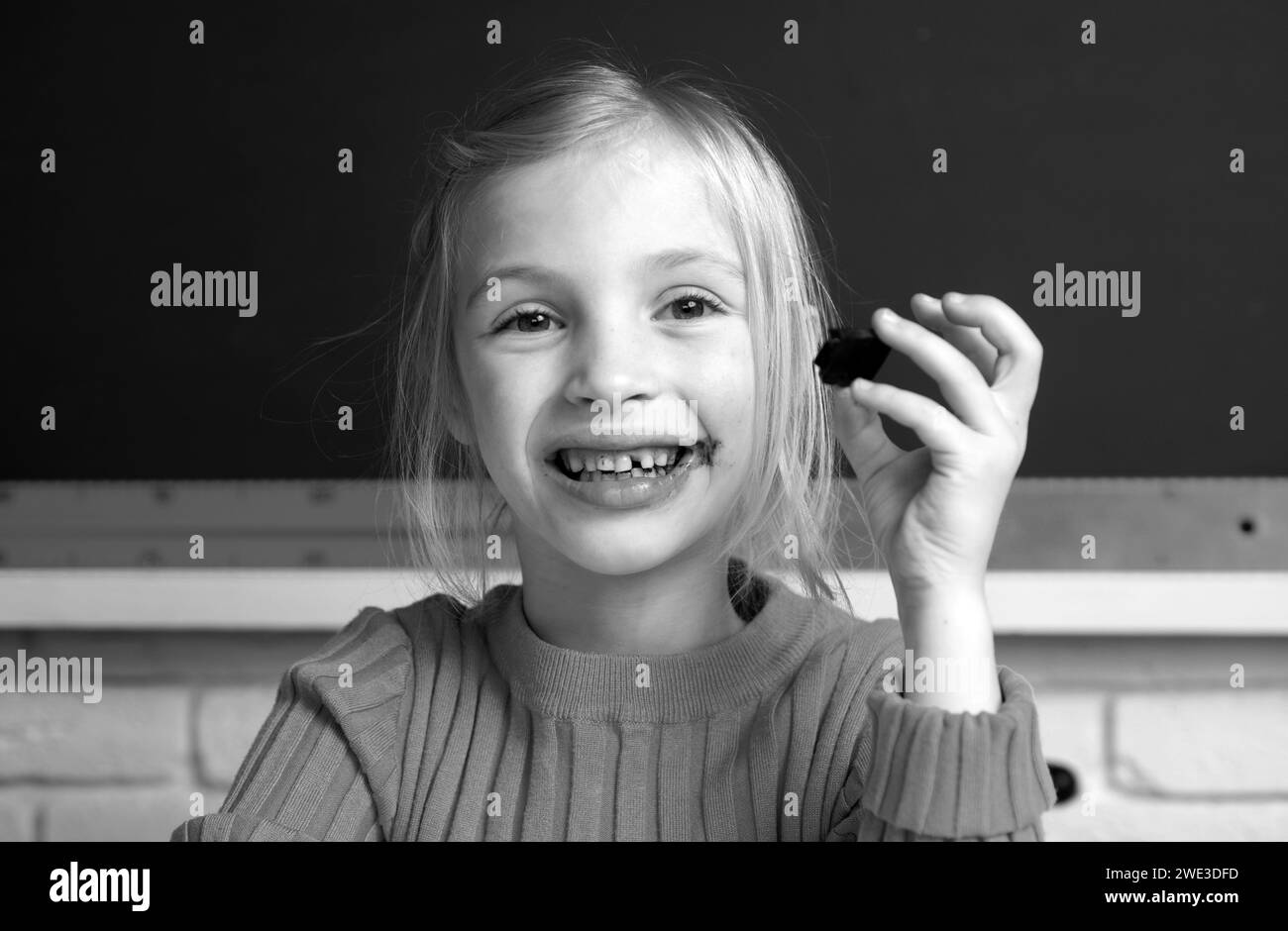 Portrait en gros plan d'une petite fille souriante assise sur un bureau de table dans la salle de classe, en train de manger du chocolat.Petite fille drôle d'école visage. Banque D'Images