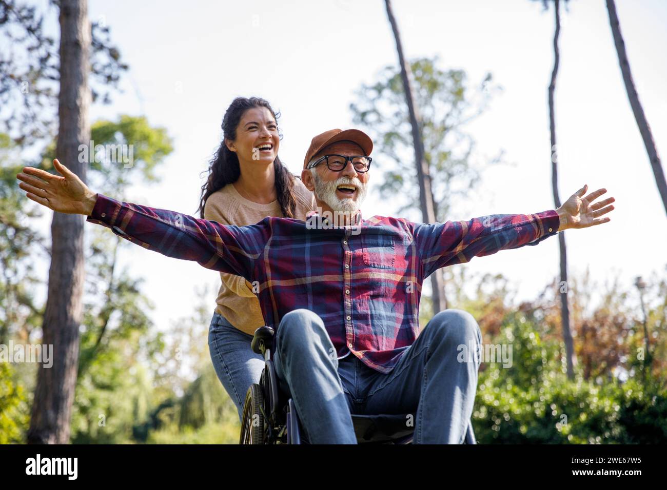 Travailleur social poussant un homme âgé retraité joyeux avec les bras tendus en fauteuil roulant au parc Banque D'Images