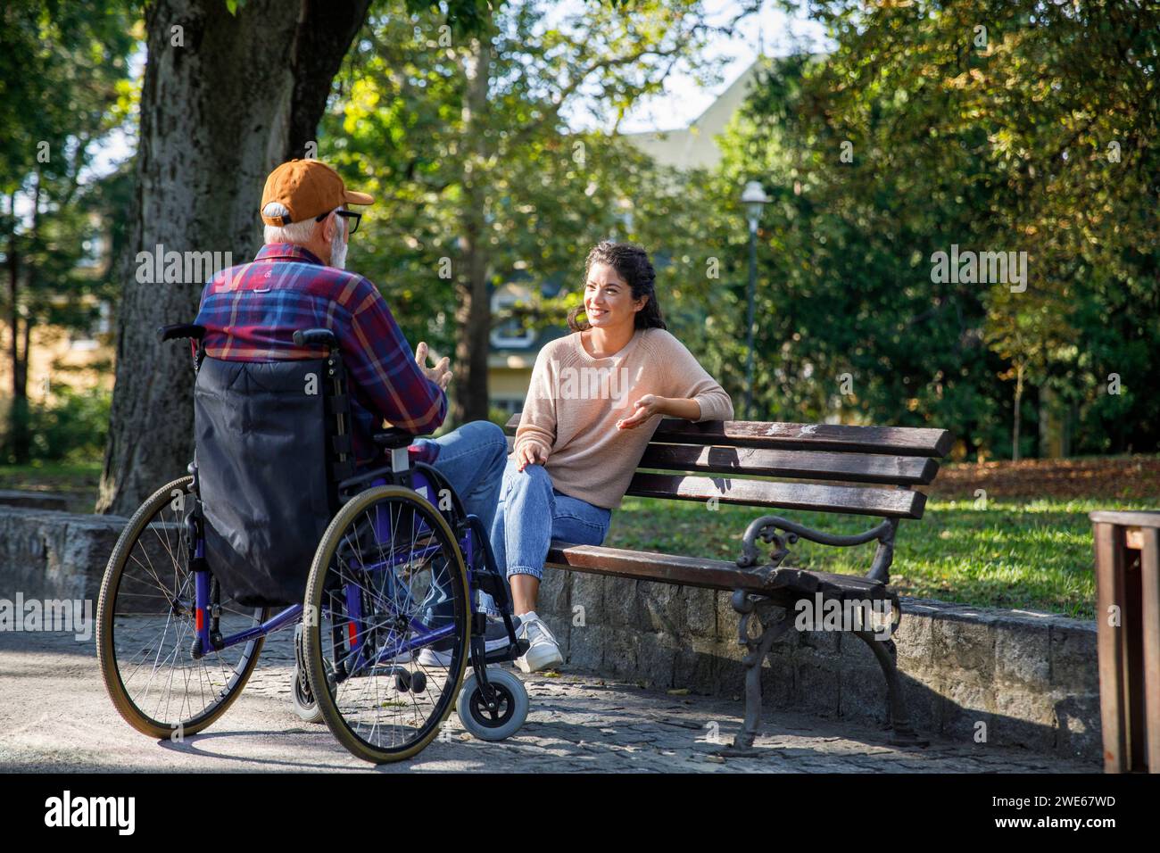 Soignant souriant assis sur un banc écoutant un homme retraité en fauteuil roulant au parc Banque D'Images