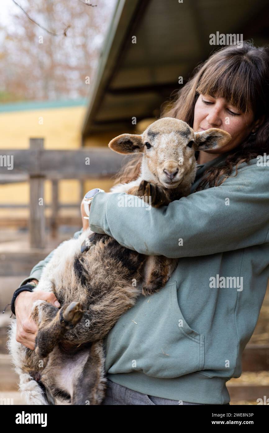 Heureuse jeune femme fermière serrant un bébé mouton sur une ferme biologique rurale. Bien-être et soins des animaux dans une grange. Banque D'Images