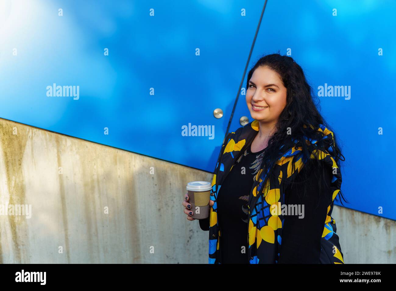 Belle jeune femme brune avec de longs cheveux bouclés, habillée d'un gilet noir, bleu et jaune, boit du café d'une tasse en papier sur un fond de Banque D'Images