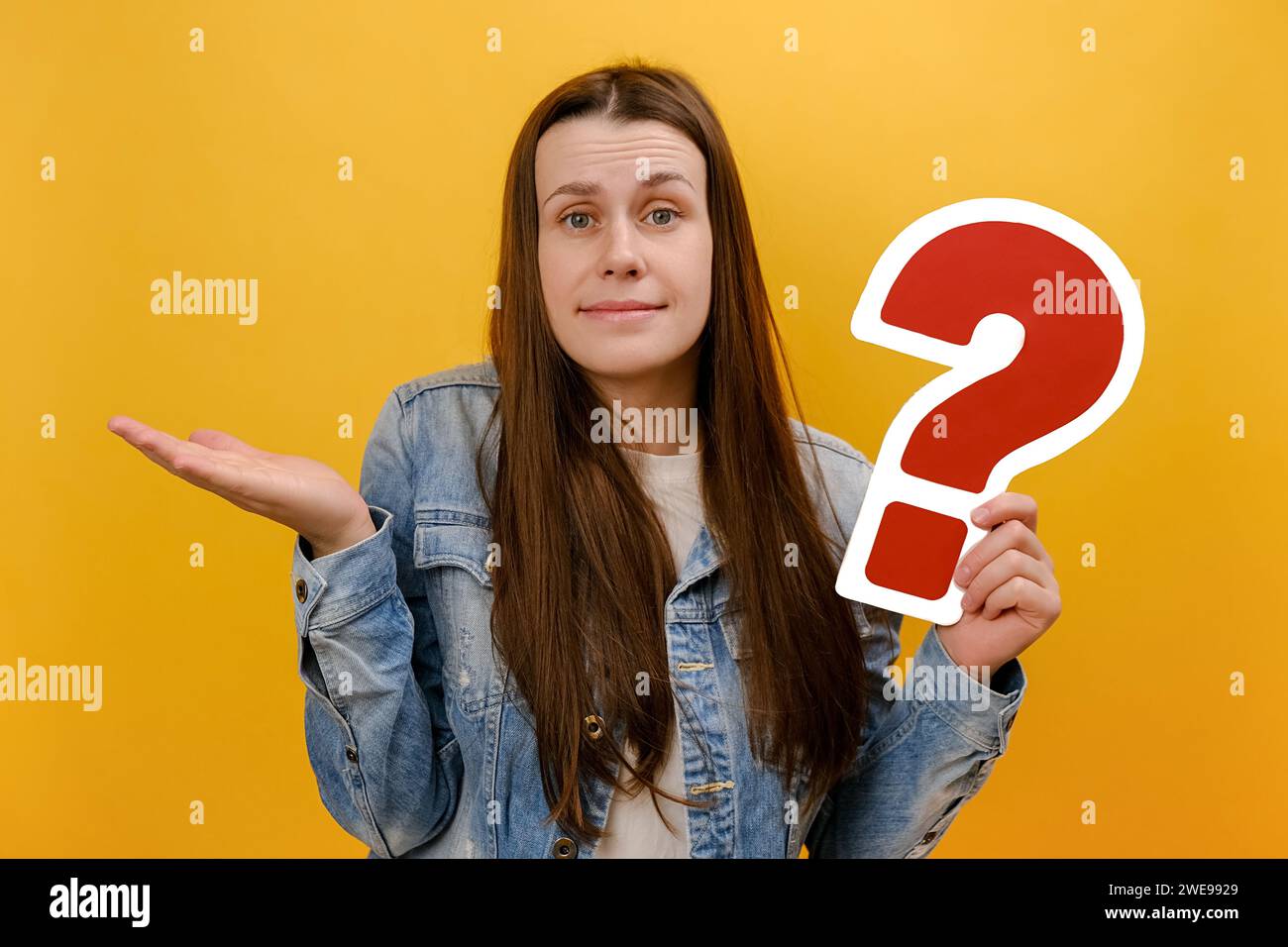 Portrait de jeune femme réfléchie confuse regardant la caméra, tenant le papier point d'interrogation rouge, pense aux tâches, portant une veste en denim, posant isola Banque D'Images