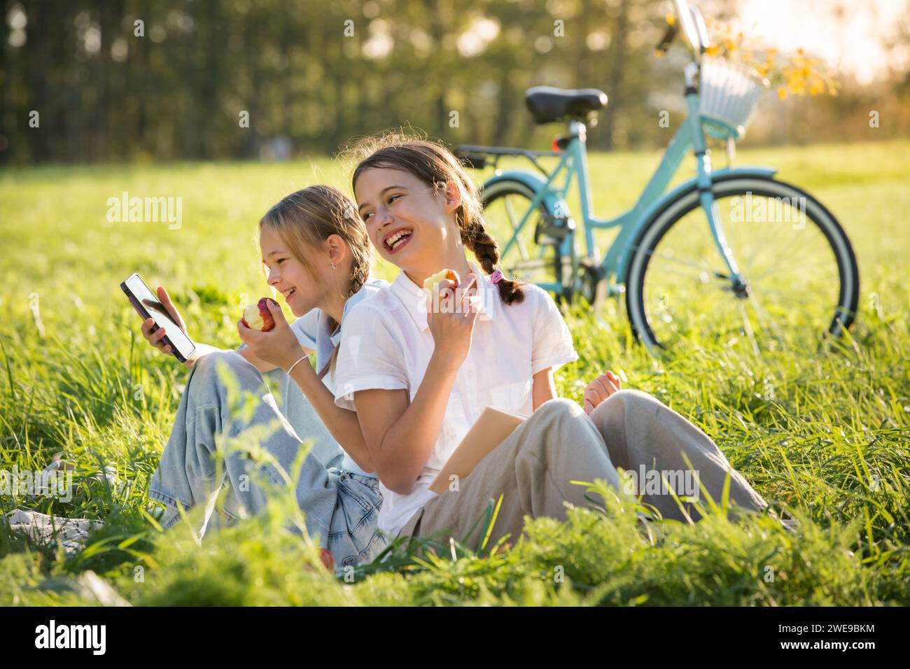 Deux filles passent du temps sur la pelouse verte dans le parc ensemble, font défiler les smartphones, lisent des livres, mangent des pommes, profitent de l'été et des vacances Banque D'Images