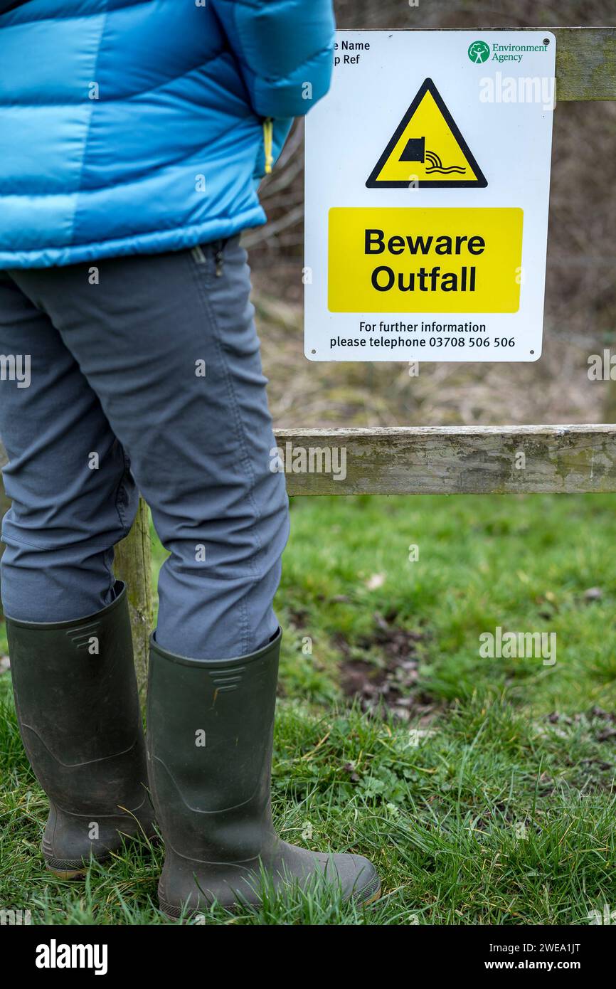 Vue arrière, angle bas, d'une personne isolée en bottes debout regardant un panneau d'avertissement de danger de l'Agence de l'environnement conseillant au public de ne pas se soucier de l'émissaire. Banque D'Images