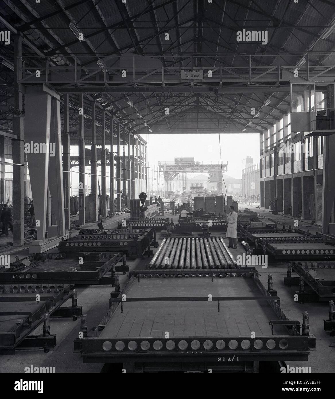 Années 1960, historique, superviseur en manteau blanc debout dans un bâtiment industriel géant de «cintre» avec portique suspendu, produisant de grands panneaux de béton prémoulés, Angleterre, Royaume-Uni. La Grande-Bretagne à la fin des années 50 et 60 a vu de grandes tours et blocs de bureaux monter, construits à partir de panneaux de béton prémoulés, ce qui signifie une construction plus rapide et moins coûteuse et une résistance au feu supérieure. Le style architectural était appelé Brutalisme. À l'intérieur de l'unité, le nom sur le portique, prêtre Rowland de Cradley Heath, Staffordshire. Ingénieurs hydrauliques britanniques, qui fabriquaient des poulies industrielles, des moufles et des chaînes. Banque D'Images