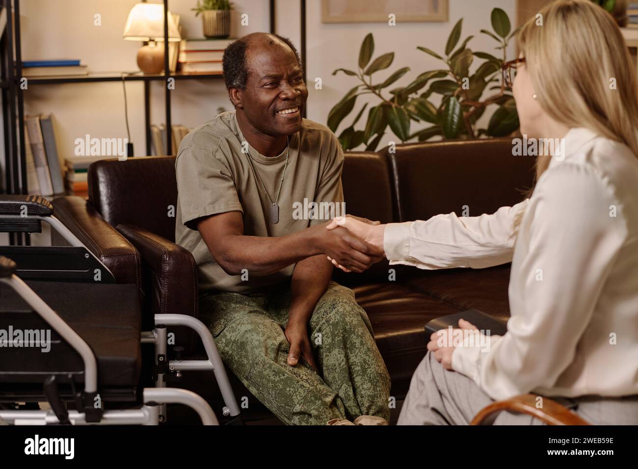 Joyeux homme noir avec handicap serrant la main avec une psychologue féminine assise sur un canapé au bureau Banque D'Images