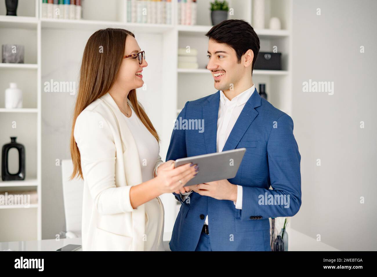 Un homme d'affaires hispanique amical s'engage dans une discussion avec sa collègue féminine sur une tablette dans un bureau lumineux et moderne, suggérant une communication efficace et une relation de travail positive Banque D'Images