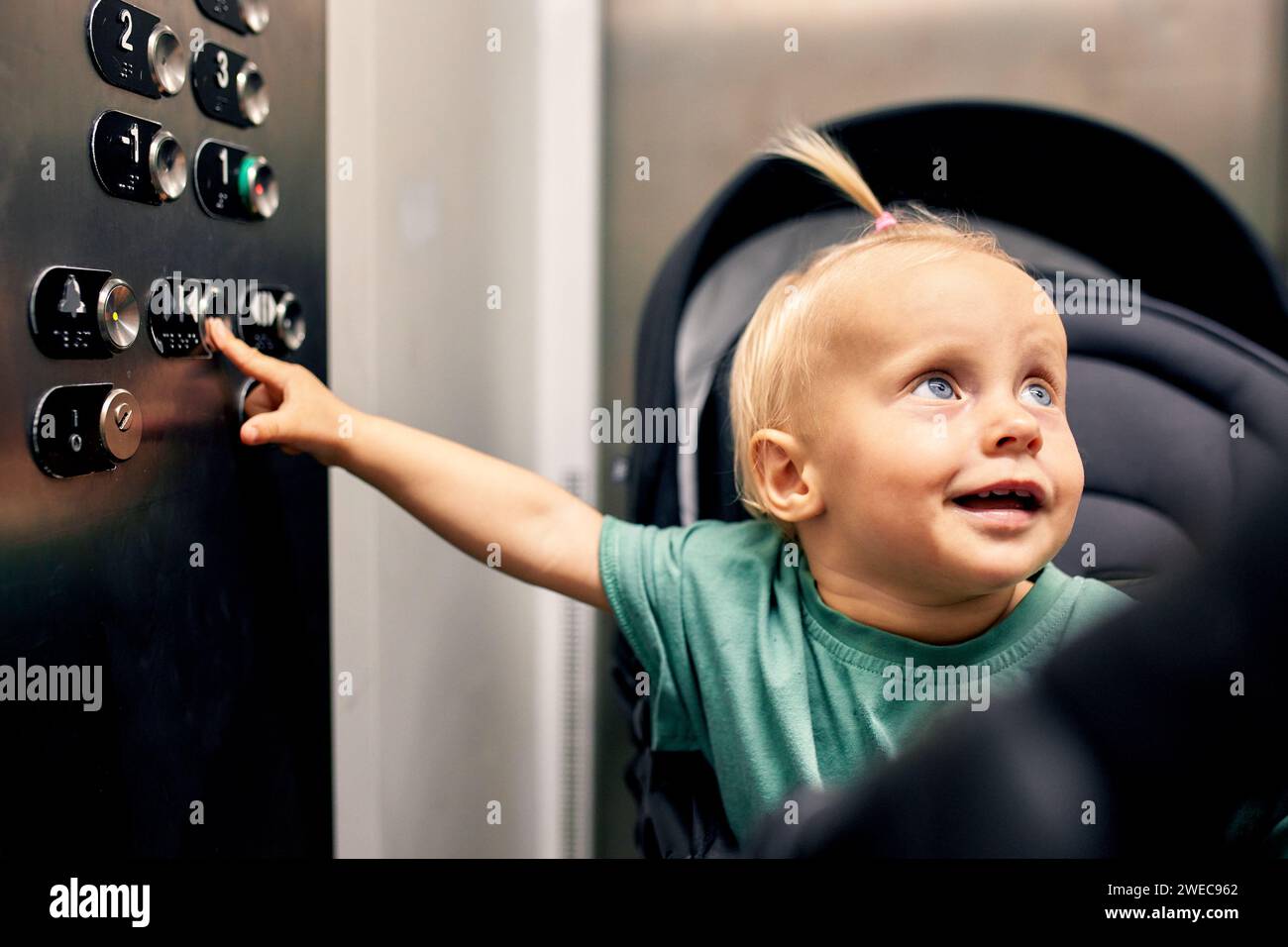 Petit bébé mignon assis dans une poussette de bébé appuie sur le bouton de plancher dans l'ascenseur. Une petite blonde dans une voiture monte ou descend l'ascenseur avec lui Banque D'Images