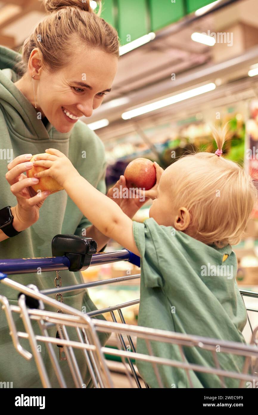 Heureuse jeune mère avec sa fille mignonne dans le chariot marchant autour du supermarché. Belle maman et petite petite fille blonde choisissant des fruits et Banque D'Images