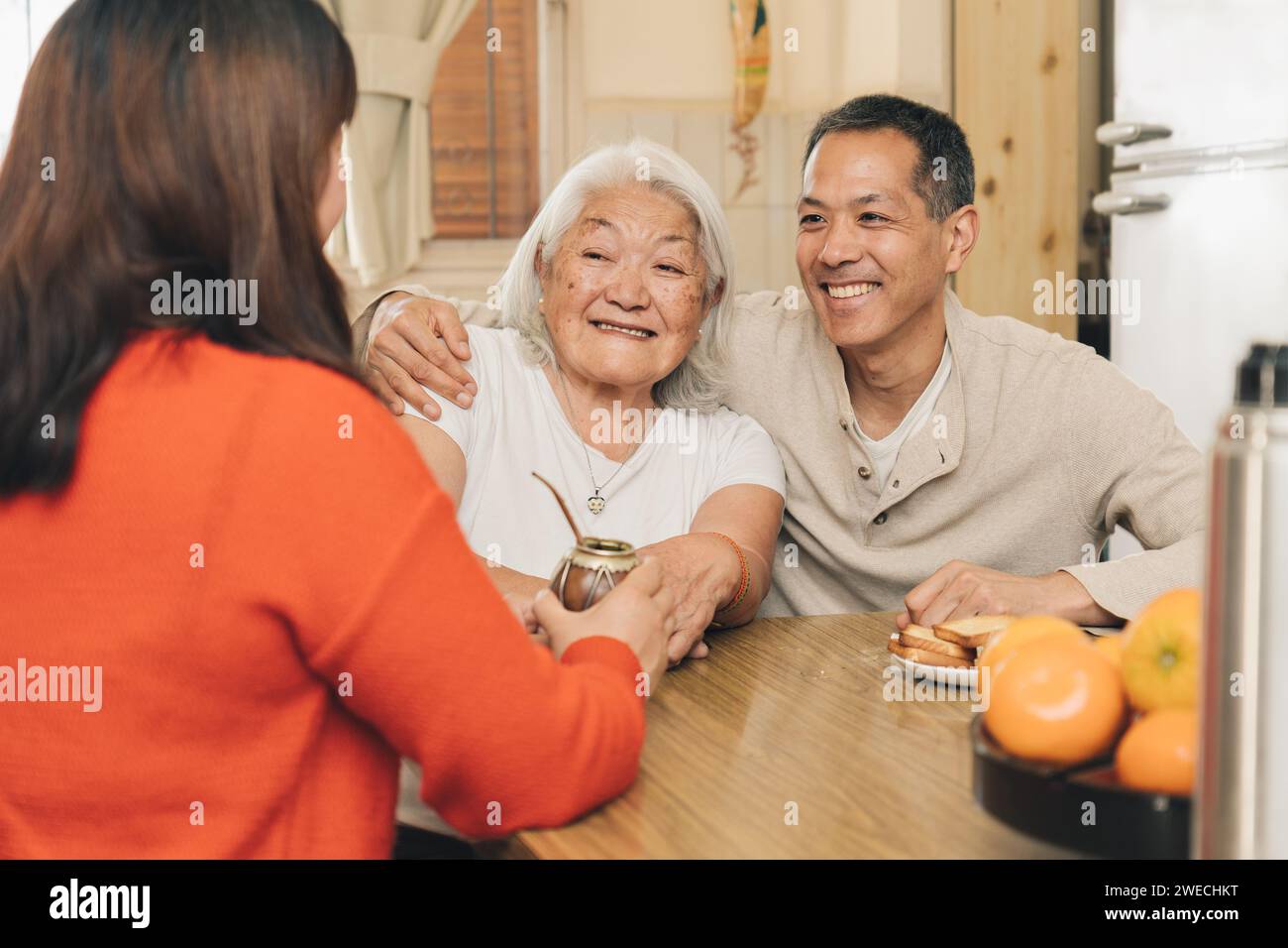 Heureux enfants adultes partageant avec leur mère japonaise âgée buvant compagnon dans la cuisine. Famille argentino-japonaise. Banque D'Images