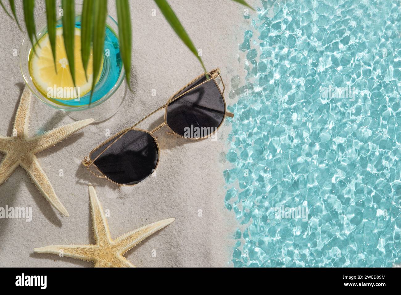 Un lac au bord du sable avec des lunettes de soleil, deux étoiles de mer et un verre à cocktail rempli de liquide bleu et une tranche de citron. Ange plat Banque D'Images