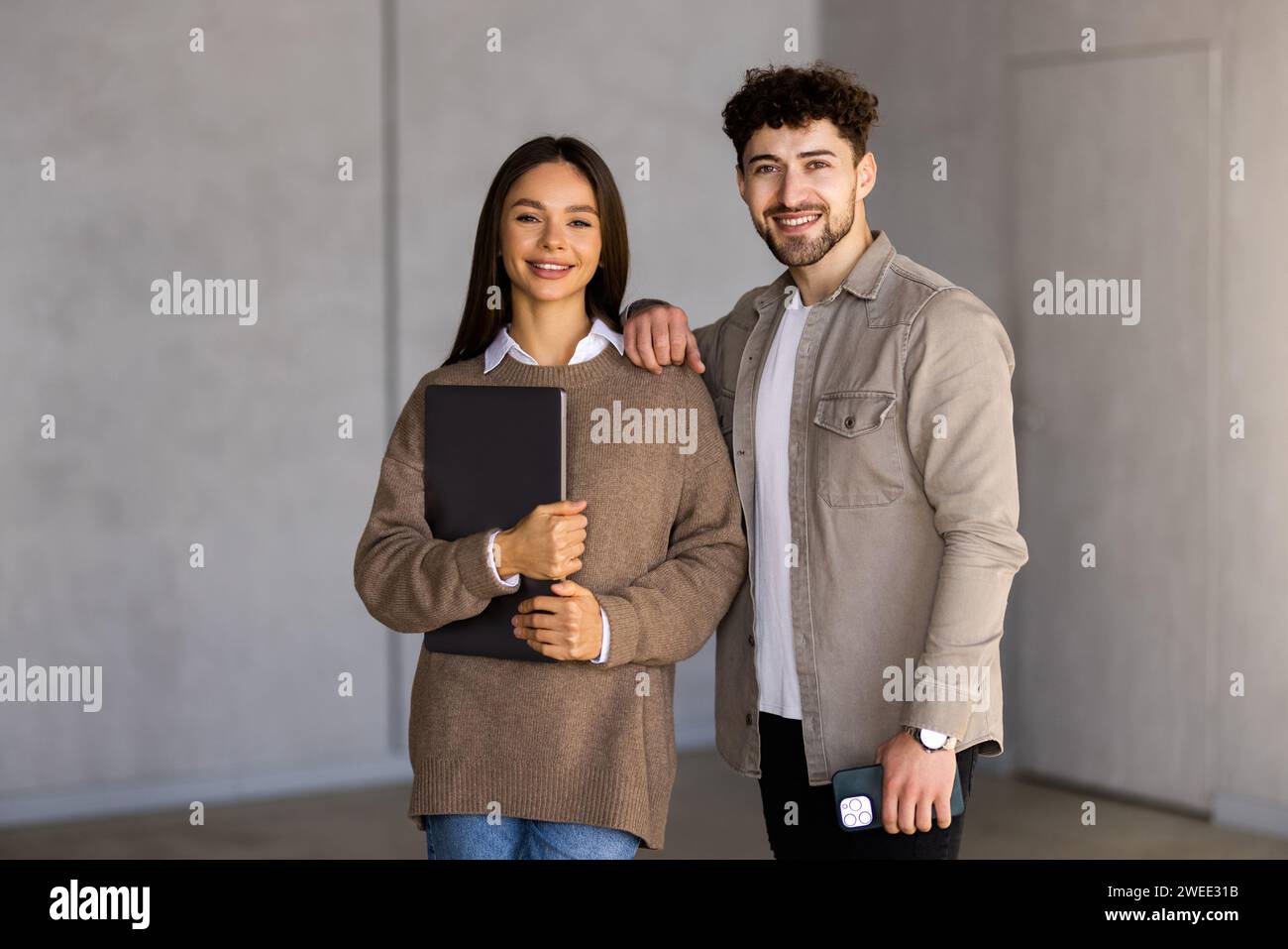 Belle jeune femme d'affaires et bel homme d'affaires iare à l'aide d'une tablette numérique, parlant et souriant, debout devant le mur gris Banque D'Images