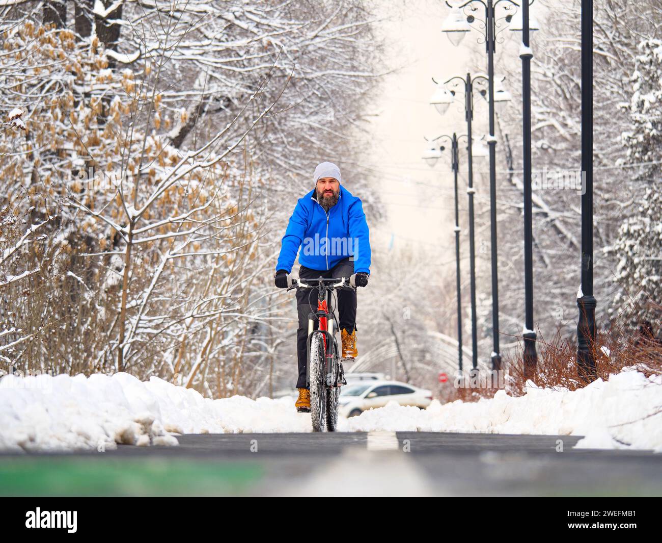 Un homme barbu fait du vélo le long d'une piste cyclable dans la ville en hiver. Mode de vie actif. Gars dans une veste bleue sur un vélo Banque D'Images