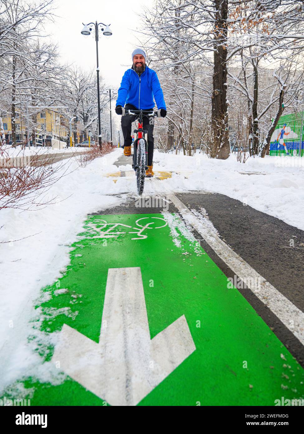 Un homme souriant fait du vélo le long d'une piste cyclable en hiver. Mode de vie actif en hiver. Homme barbu dans une veste bleue sur un vélo rouge Banque D'Images