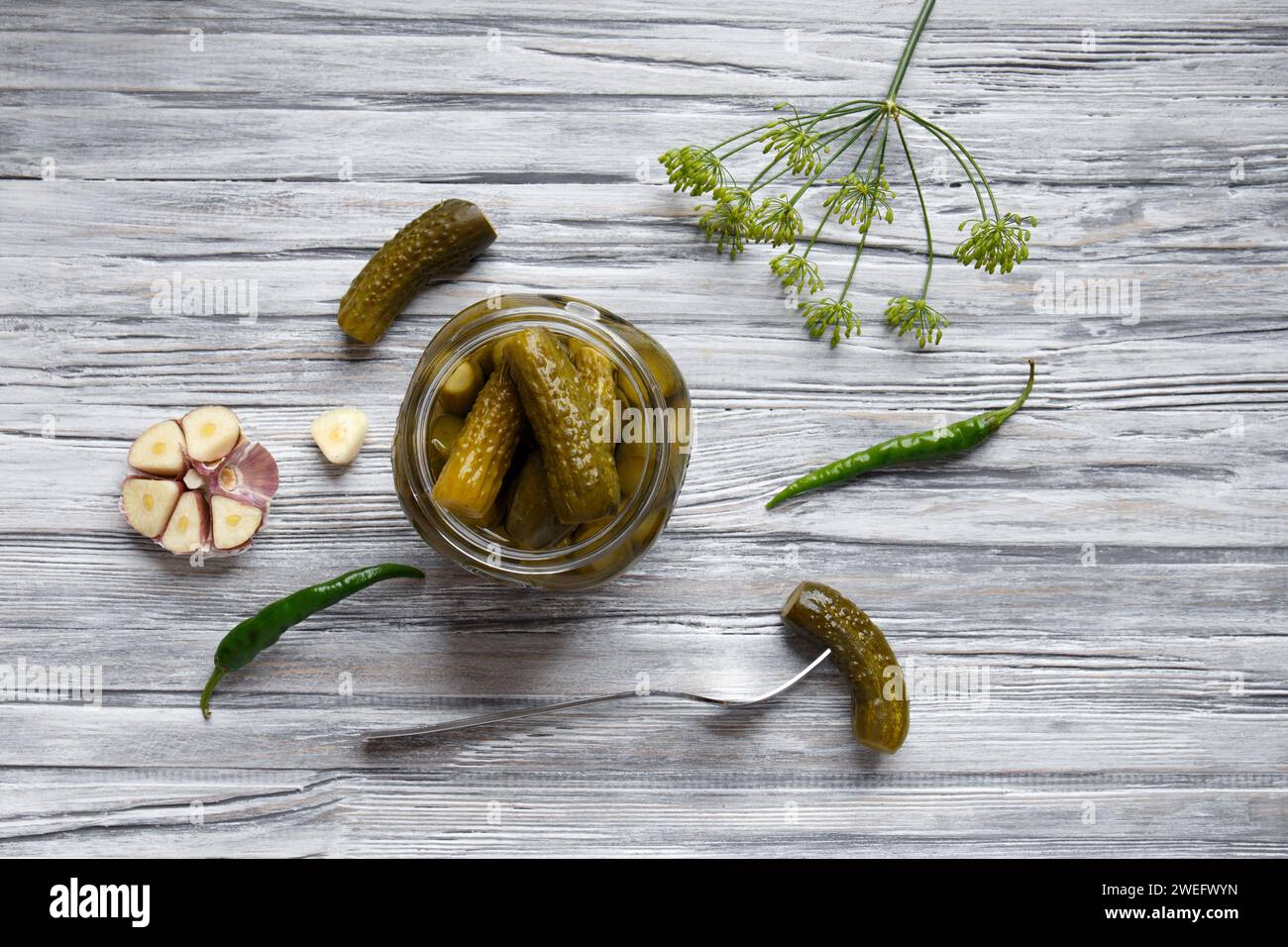 Concombres en conserve dans un bocal ouvert, un cornichon mariné est piqué sur une fourchette et se trouve à proximité sur un fond en bois gris, vue de dessus. Banque D'Images