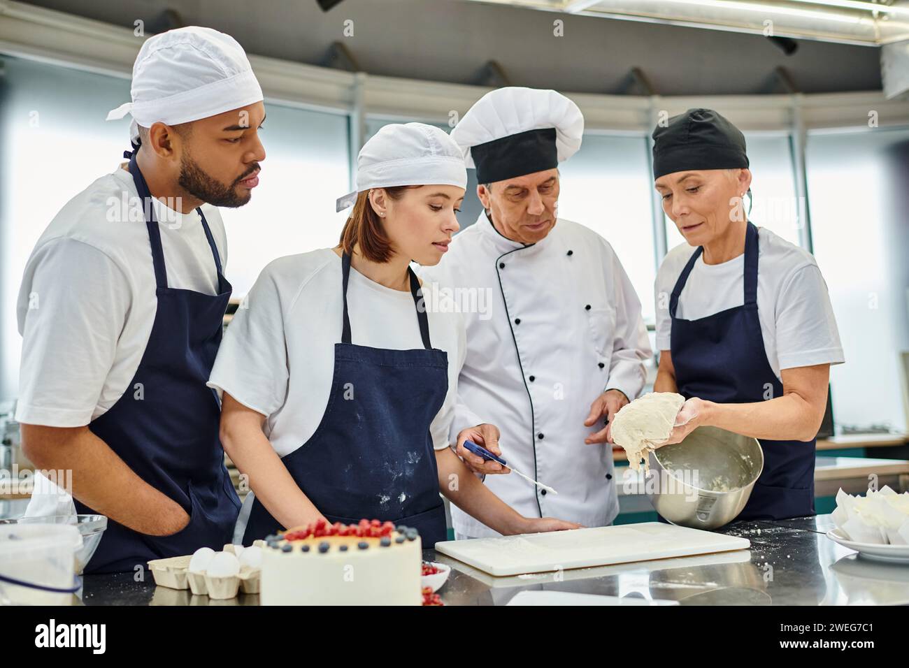 beau chef cuisinier mature en chapeau blanc regardant comment ses divers chefs en tabliers travaillent avec de la pâte Banque D'Images