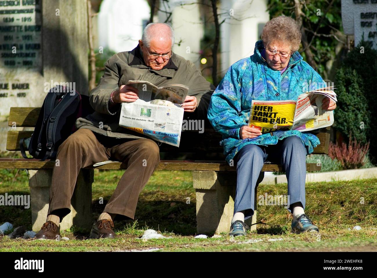 Un couple de personnes âgées est assis sur un banc sous le soleil du printemps lisant un journal, les mots «ici l'ours danse» peuvent être vus Banque D'Images