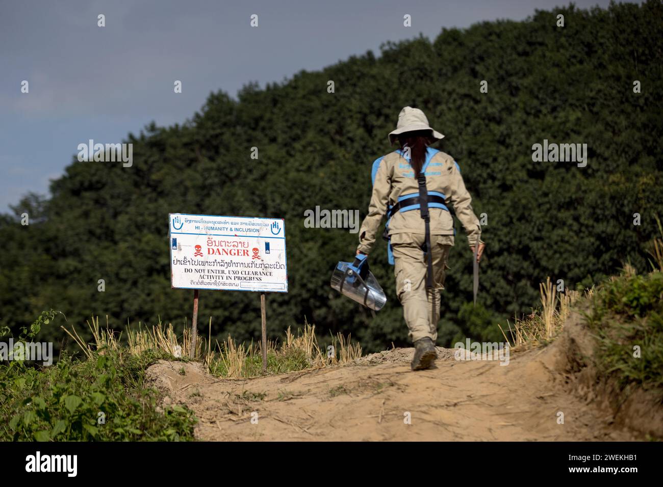 Un opérateur sur le terrain portant un équipement de protection individuelle passe devant un panneau ÔDangerÕ près de Muang Mai, Laos, le 25 octobre 2023 à côté d'un riz parsemé de munitions non explosées (UXO) qui restent enfouies dans le sol après la campagne de bombardement des États-Unis pendant la guerre du Vietnam. Le site est nettoyé par des opérateurs de terrain ou des démineurs travaillant pour l'organisation non gouvernementale internationale humanité et inclusion. Banque D'Images