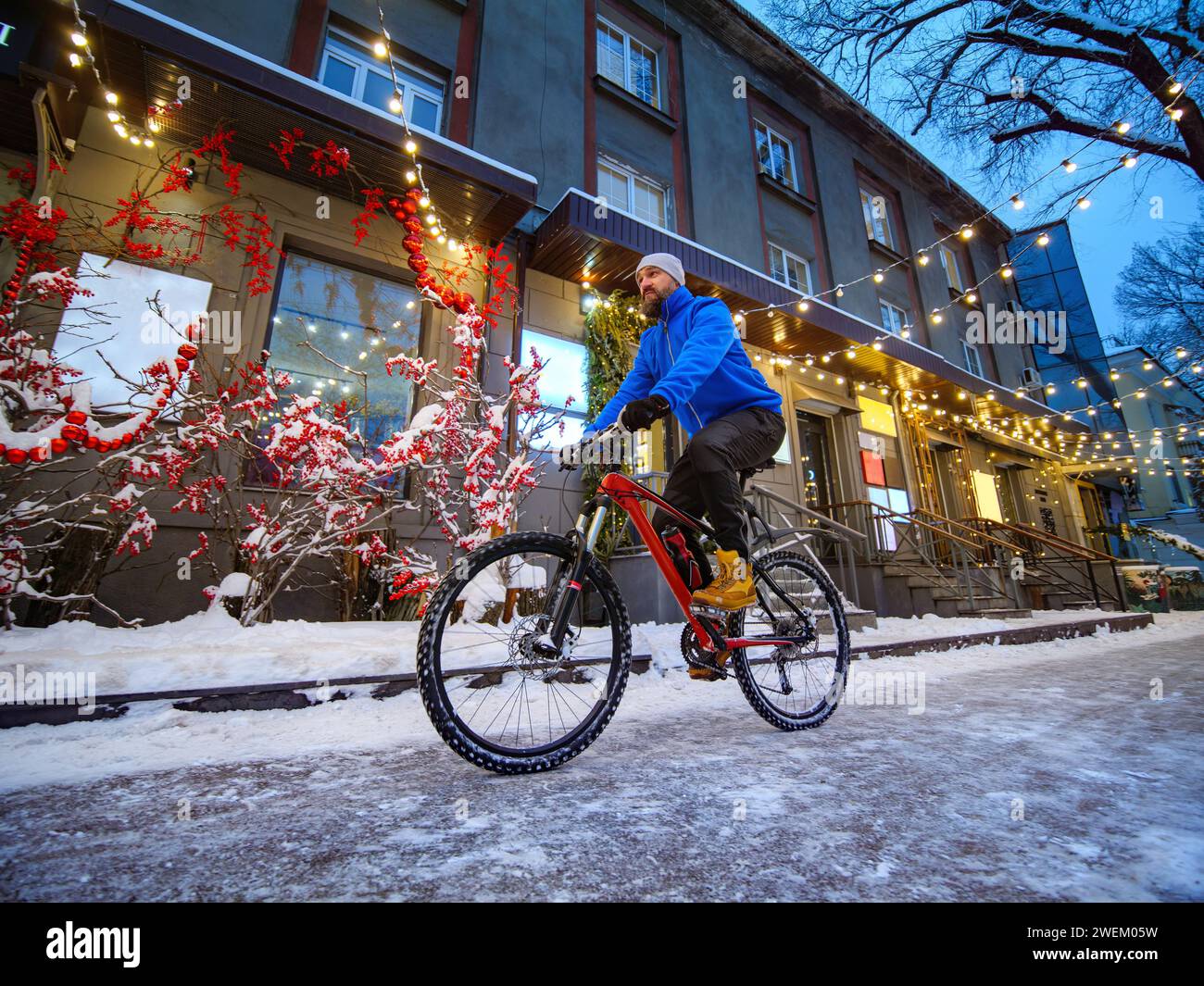 Un homme monte à vélo le long d'une rue joliment décorée dans une ville d'hiver. Homme barbu dans une veste bleue sur un vélo rouge. Ville à Noël Banque D'Images