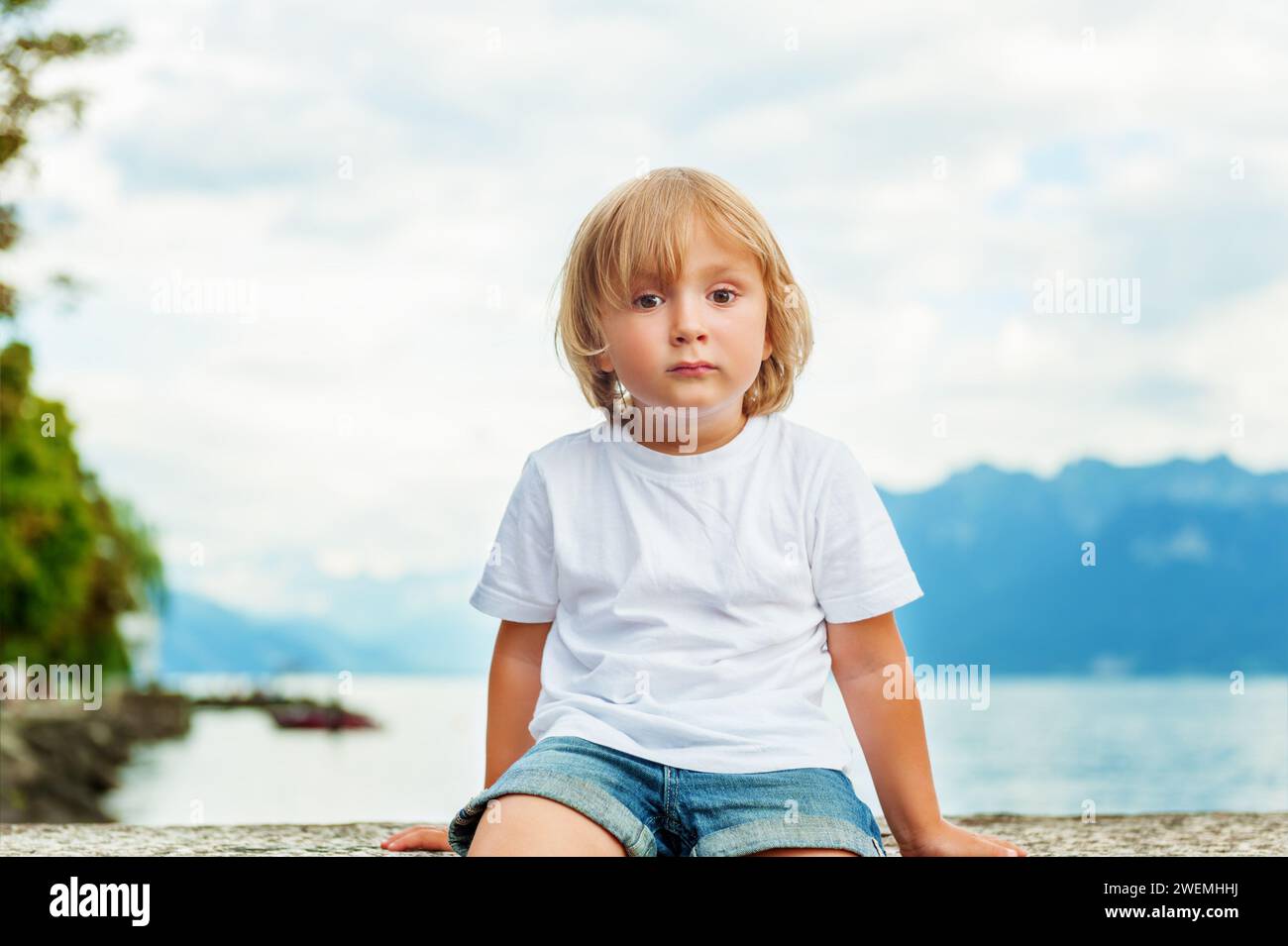 Portrait de beau souriant mignon petit garçon. Petit enfant de 3-4 ans jouant dehors au bord du lac Léman, en été ou au printemps. Garçon assis seul b Banque D'Images