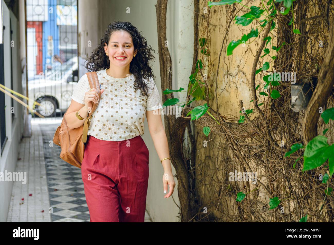 Jeune femme latina vénézuélienne élégante, en curleurs, avec sac à dos, souriant heureux dans le couloir de la résidence arrivant à la maison, concept de style de vie avec spa copie Banque D'Images