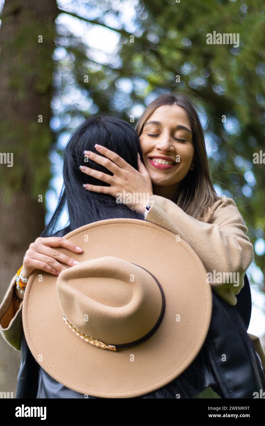 Image verticale d'une femme heureuse serrant son amie les yeux fermés dans le parc tout en tenant son chapeau Banque D'Images