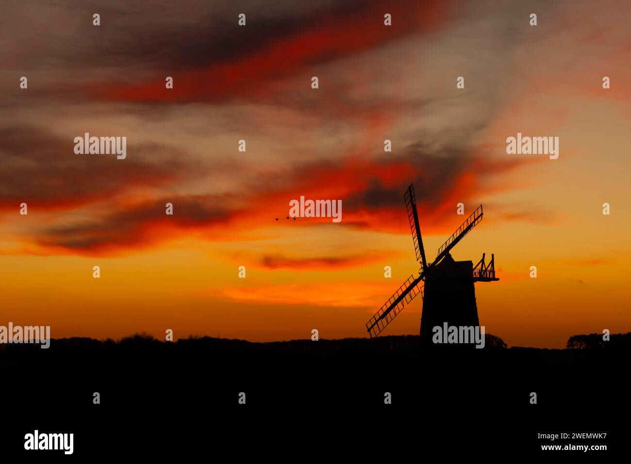 Moulin à vent silhouette au coucher du soleil avec un ciel rouge et des nuages et trois oies à pieds roses (Anser brachyrhynchus) volant au-dessus, Burnham Ovary Staithe Banque D'Images