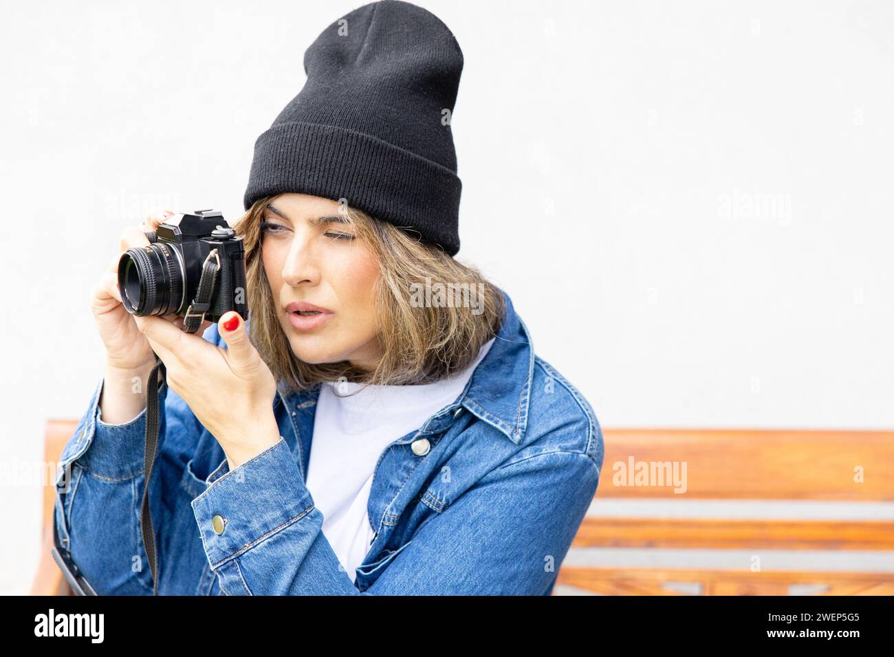 Une photographe engagée avec un appareil photo vintage se concentre intensément sur son sujet, habillée dans un style urbain décontracté avec une veste en denim et un bonnet. Banque D'Images