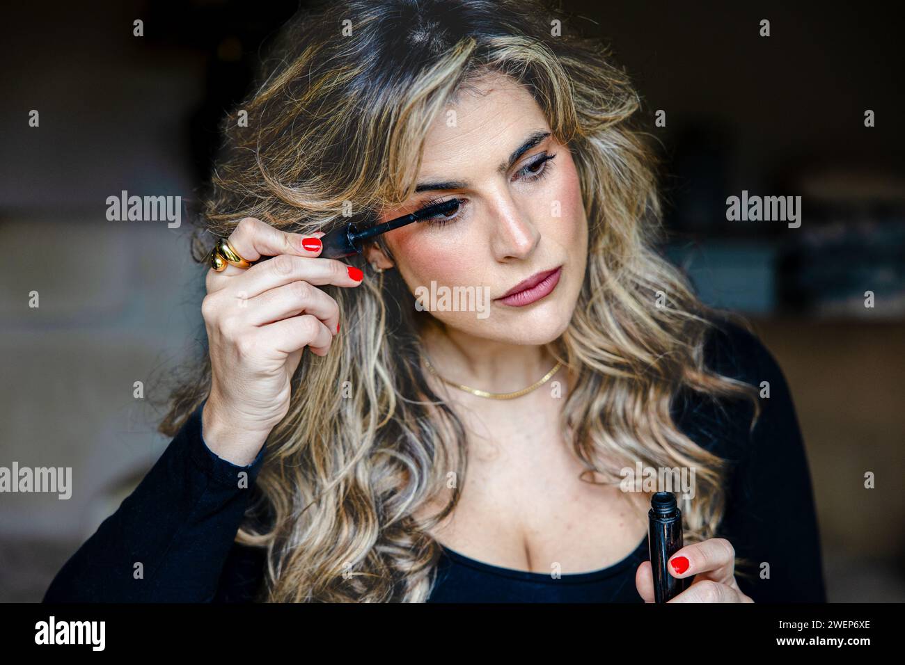 Femme élégante avec des boucles mises en évidence applique méticuleusement le mascara, améliorant sa beauté naturelle dans son cadre à la maison. Banque D'Images