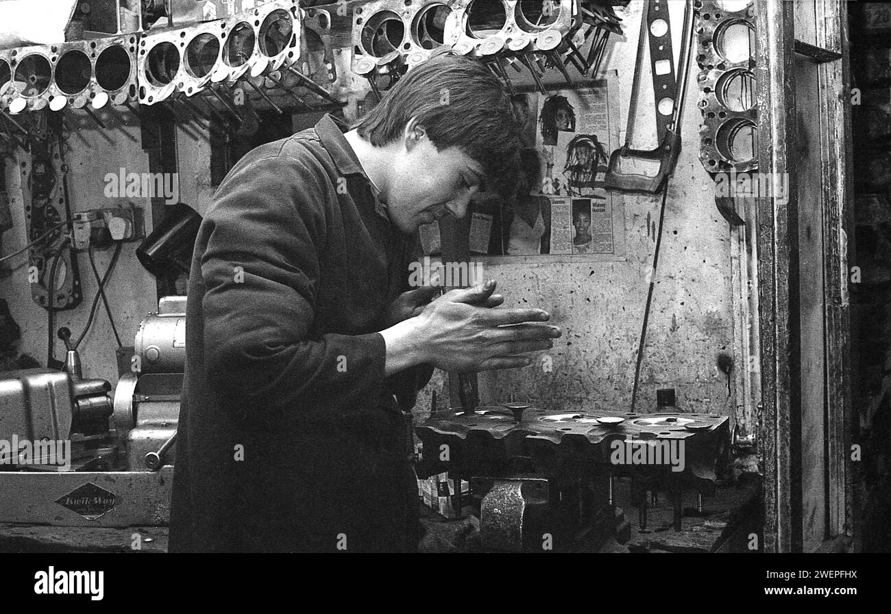 Années 1980, historique, un jeune homme en salopette, un mécanicien automobile dans un atelier, utilisant un outil à main pour réparer la tête de cylindre d'un moteur, Angleterre, Royaume-Uni. Accrochée au mur, une sélection de cercueils de moteur, qui sont des joints entre deux surfaces de moteur. Banque D'Images