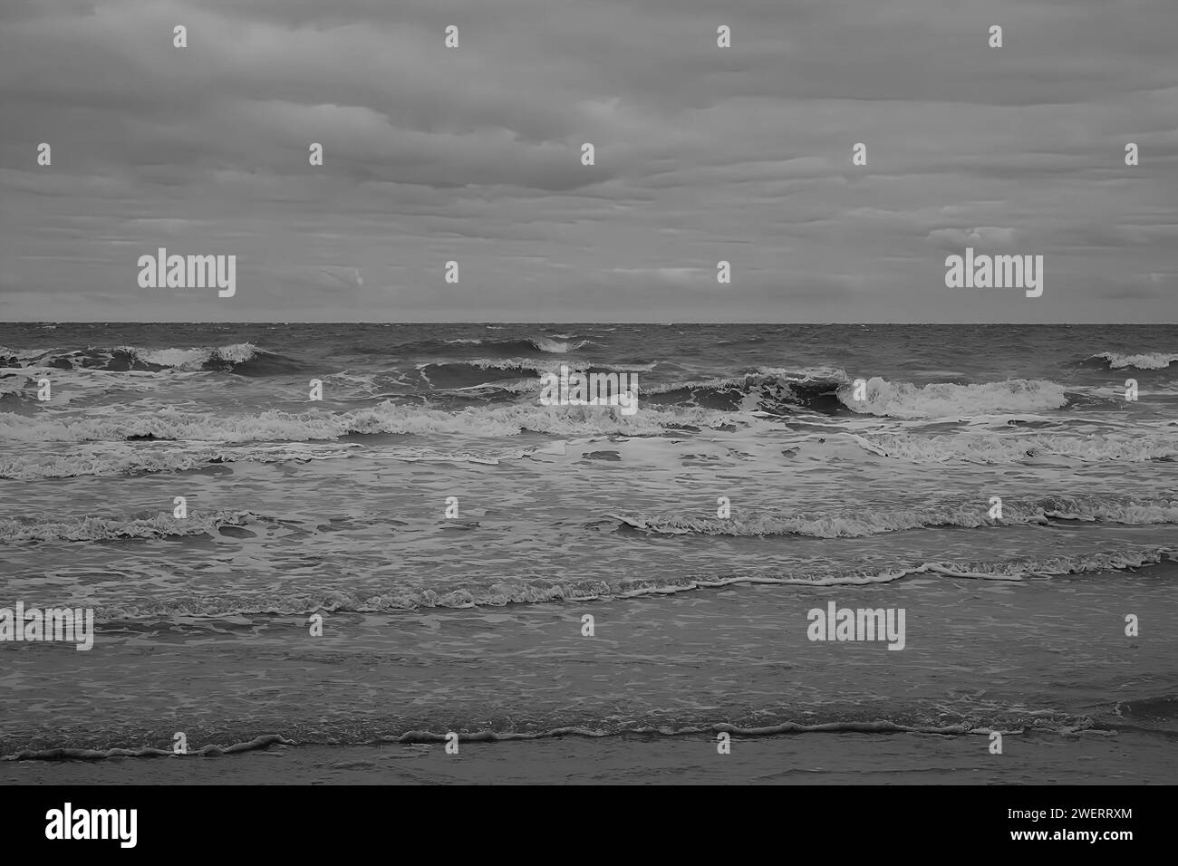 Les vagues s'écrasent sur la plage créant de l'écume blanche sous les nuages d'un ciel sombre en monochrome. Banque D'Images