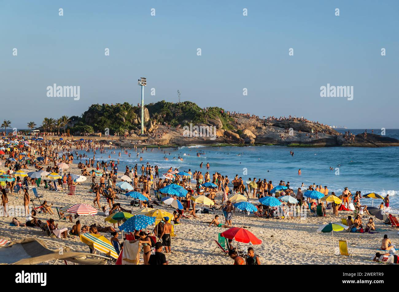 Le rivage de la plage Arpoador entièrement bondé avec Arpoador Rock (Pedra do Arpoador) péninsule à l'arrière sous l'été fin d'après-midi ciel bleu clair. Banque D'Images