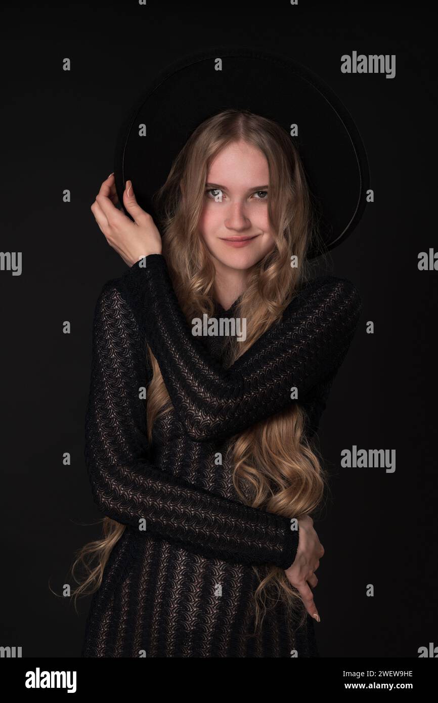 Portrait d'une jeune femme blonde jouée avec un sourire sur les lèvres, tenant gracieusement le bord de son chapeau d'une main, plaçant l'autre sur sa taille Banque D'Images